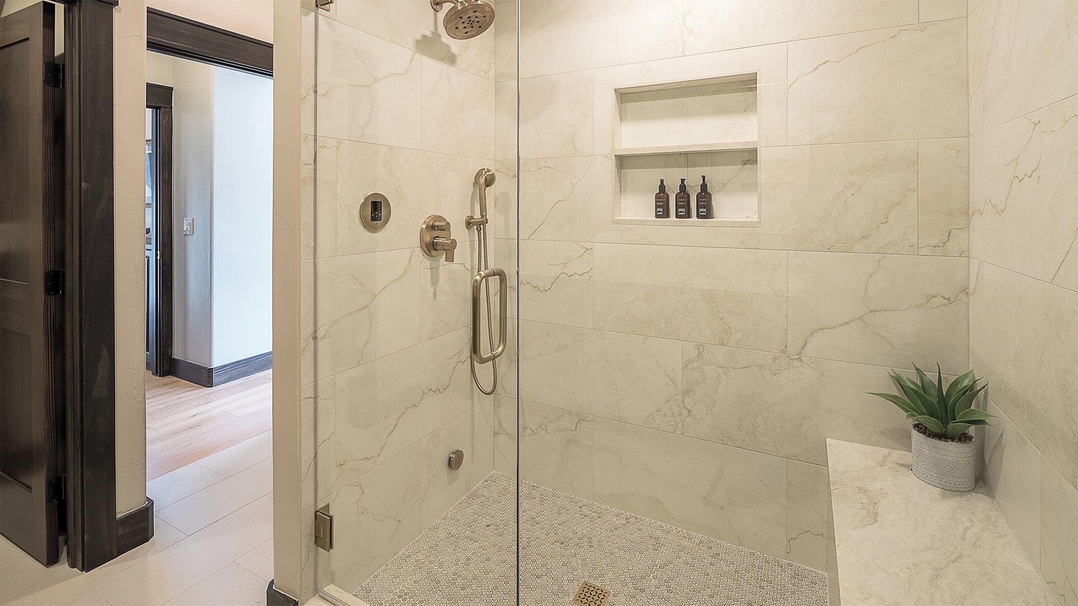 A walk-in shower with a glass door, marble walls, a small shelf with toiletries, and a potted plant.
