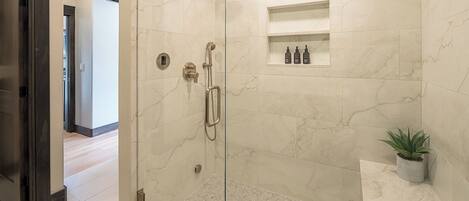 A walk-in shower with a glass door, marble walls, a small shelf with toiletries, and a potted plant.