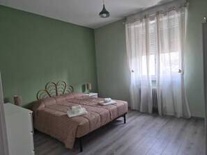 Quarto com cama, mesa de cabeceira, abajur e janela com cortinas.