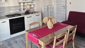 Uma cozinha moderna com fogão a gás, forno e armários brancos. Há uma mesa de jantar com cadeiras e um jogo americano vermelho. Um sofá vermelho é visível ao fundo.