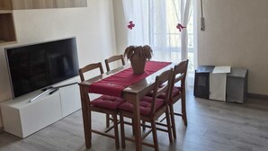 Uma sala de estar com televisão, mesa de jantar de madeira, cadeiras e um sofá vermelho.