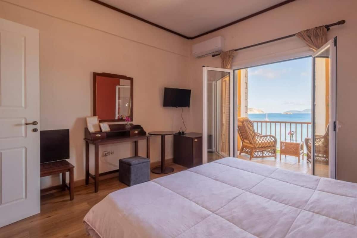 A hotel room with a bed, a desk with a flat-screen TV, a small table, and a balcony with wicker chairs.