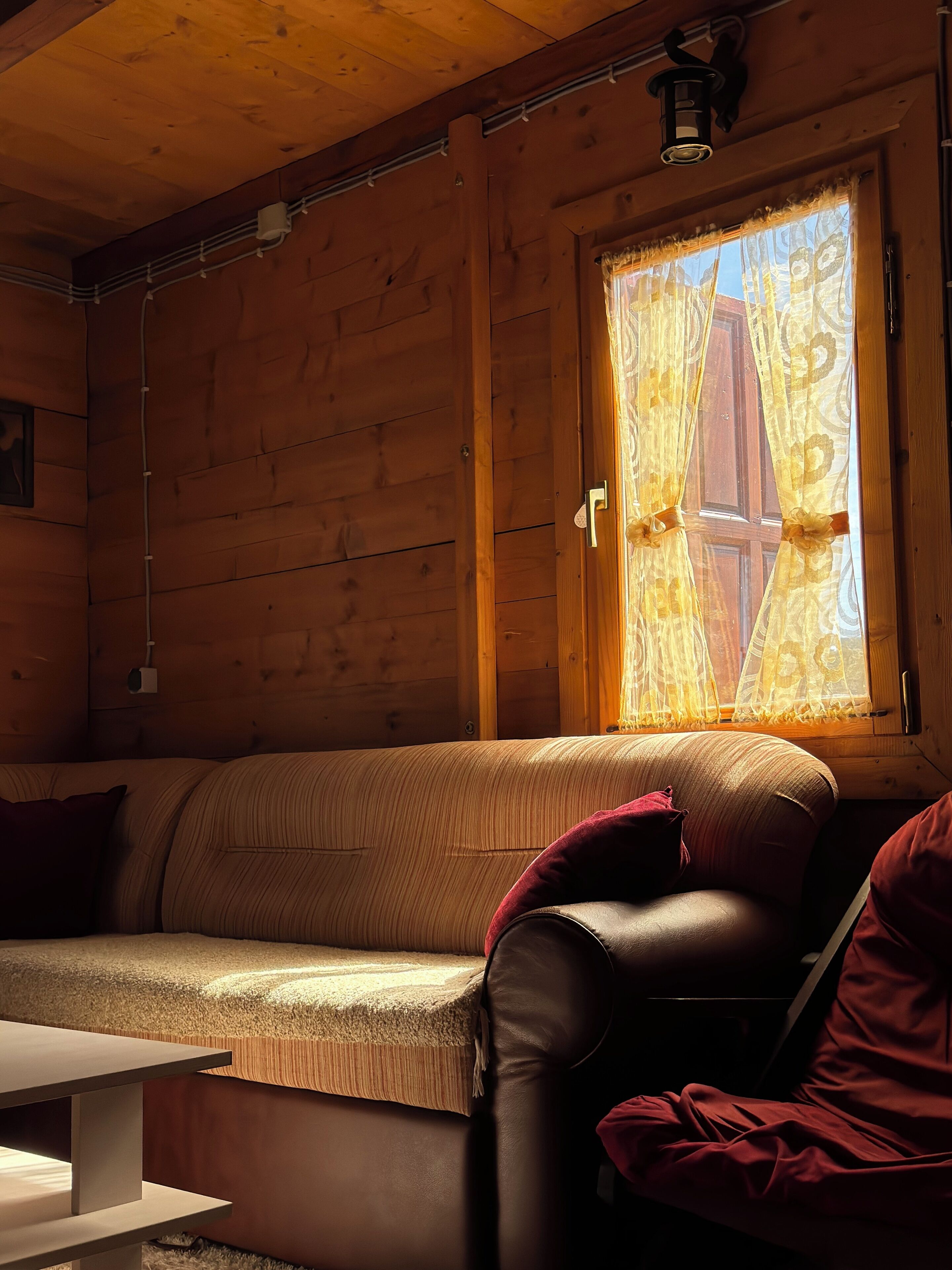 A cozy cabin interior with a wooden wall, a window with curtains, a sofa, and a table.