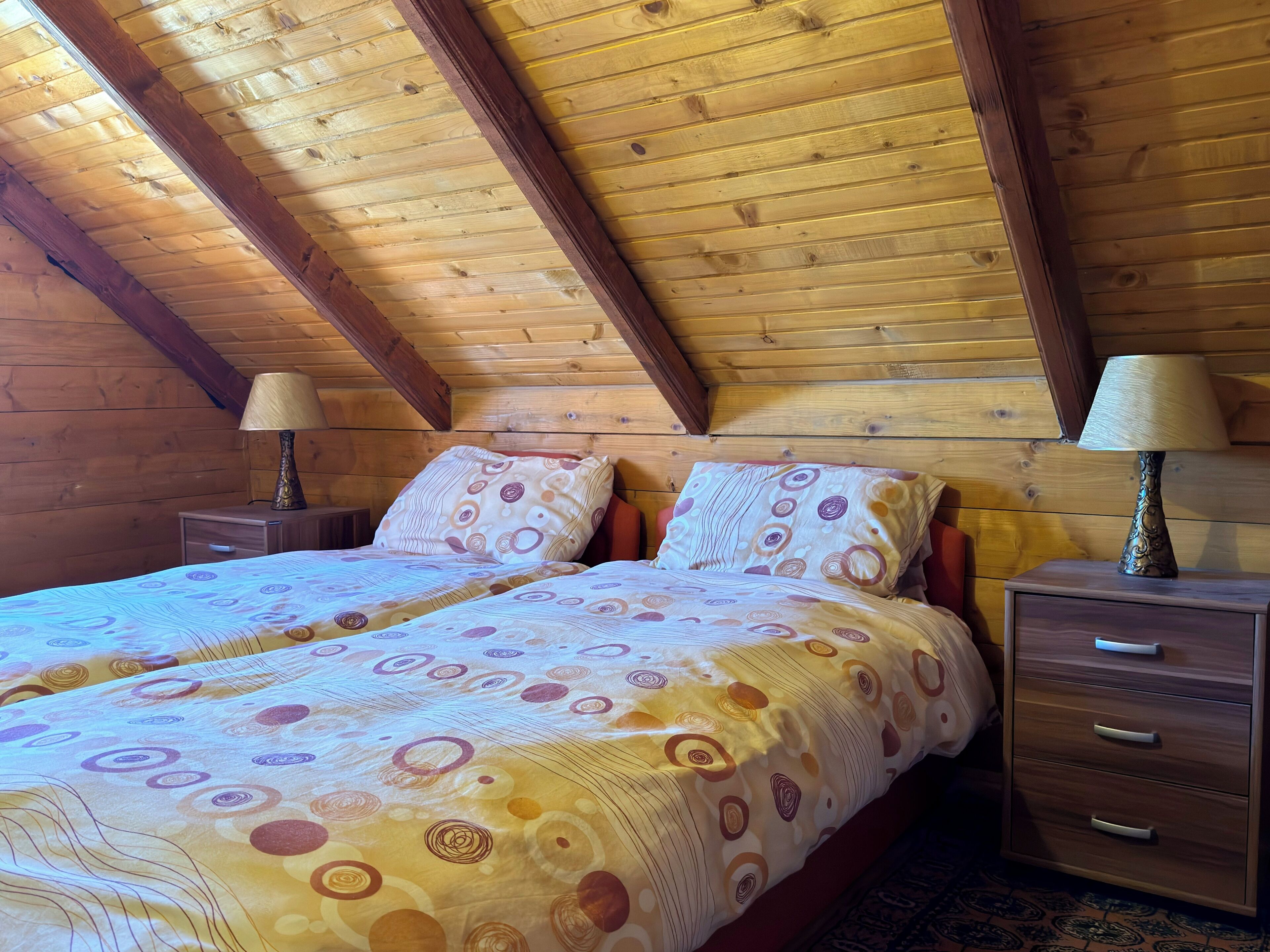 A bedroom with a sloped ceiling, wooden walls, a double bed with patterned bedding, two bedside tables with lamps, and a small drawer unit.