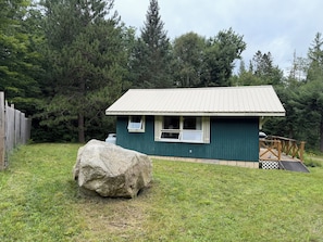 A small, single-story cabin with a metal roof and a deck, surrounded by a grassy yard and dense trees. - Cozy 1-bedroom cabin in quaint Coplin, with AC and gas fireplace. (Coplin plantation)