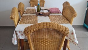 Un coin repas avec des chaises en osier, une table recouverte d’une nappe blanche, une bouteille de vin et un tableau encadré accroché au mur.