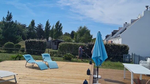 Une terrasse en bois avec des chaises longues et une table, entourée de verdure et sous un ciel dégagé.