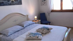 Quarto com cama, mesa de cabeceira, luminária, cadeira, janela com cortinas, ar condicionado e decoração na parede.