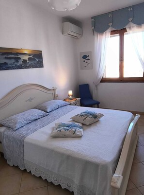 A bedroom with a bed, a nightstand, a lamp, a chair, a window with curtains, an air conditioner, and wall art. - Casa MARES, an oasis of relaxation and comfort ten minutes from the center of Olbia (Olbia)
