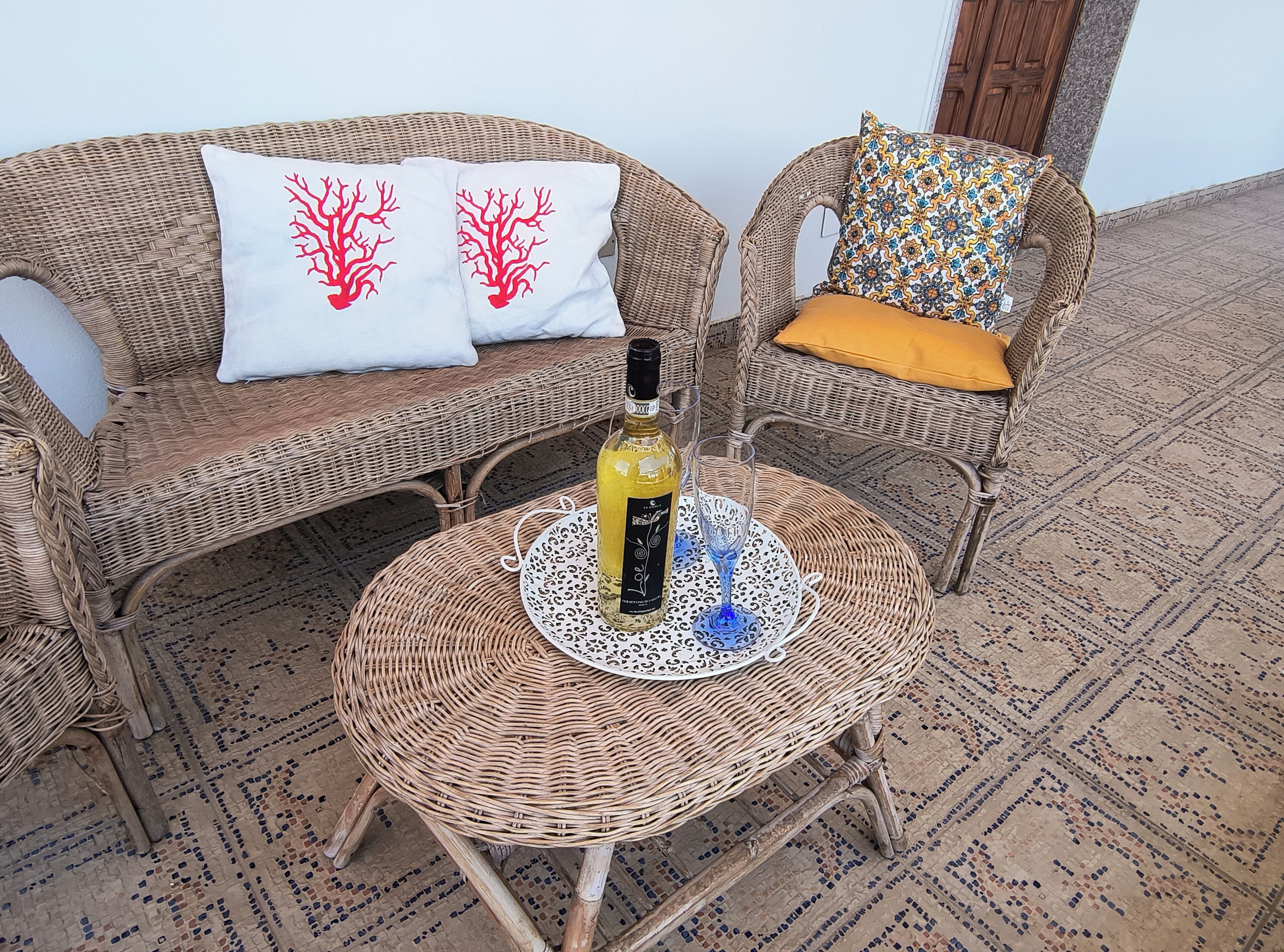 Mobili in vimini con cuscini decorati, una bottiglia di vino e un bicchiere su un tavolino intrecciato.