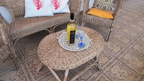 Móveis de vime com almofadas estampadas, uma garrafa de vinho e um copo sobre uma mesa trançada.