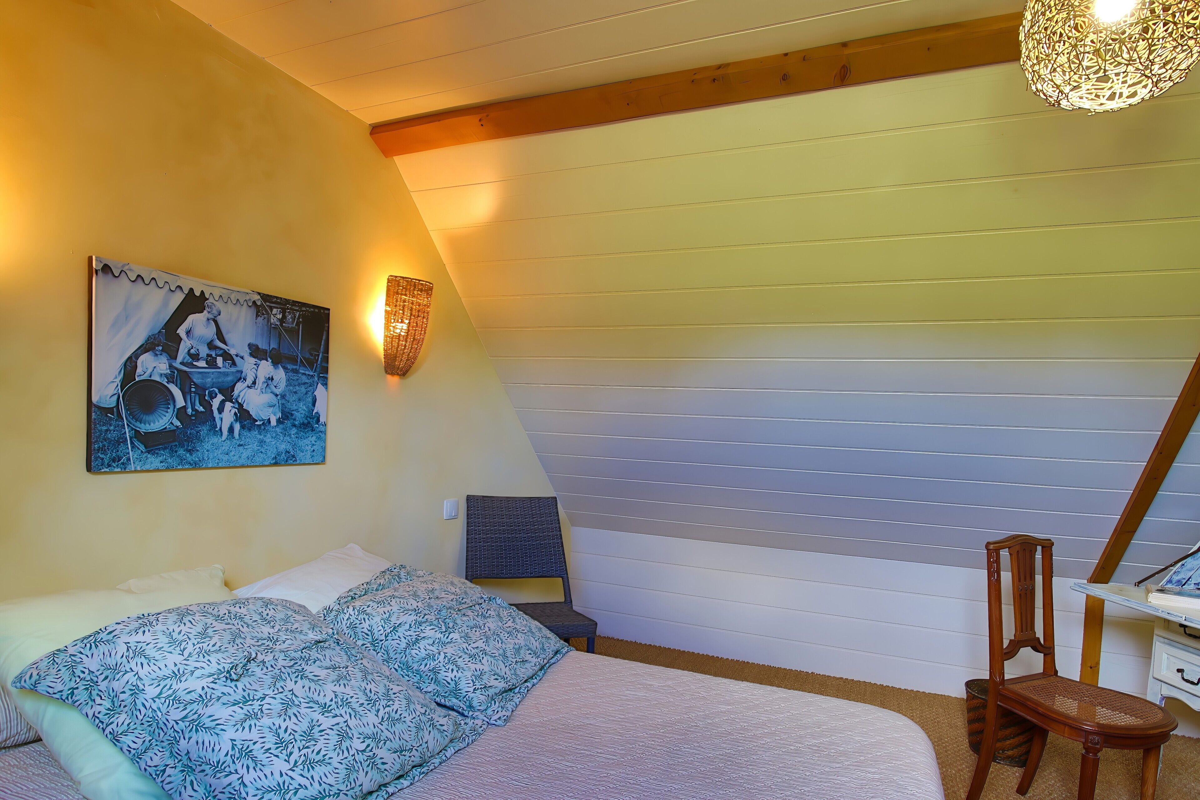 Een slaapkamer met een bed, een stoel, een bureau, een lamp en een schilderij aan de muur.
