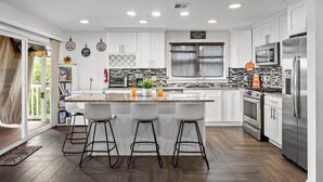 Une cuisine moderne avec des meubles blancs, un grand îlot central doté de tabourets, un réfrigérateur en acier inoxydable et une fenêtre avec des stores.