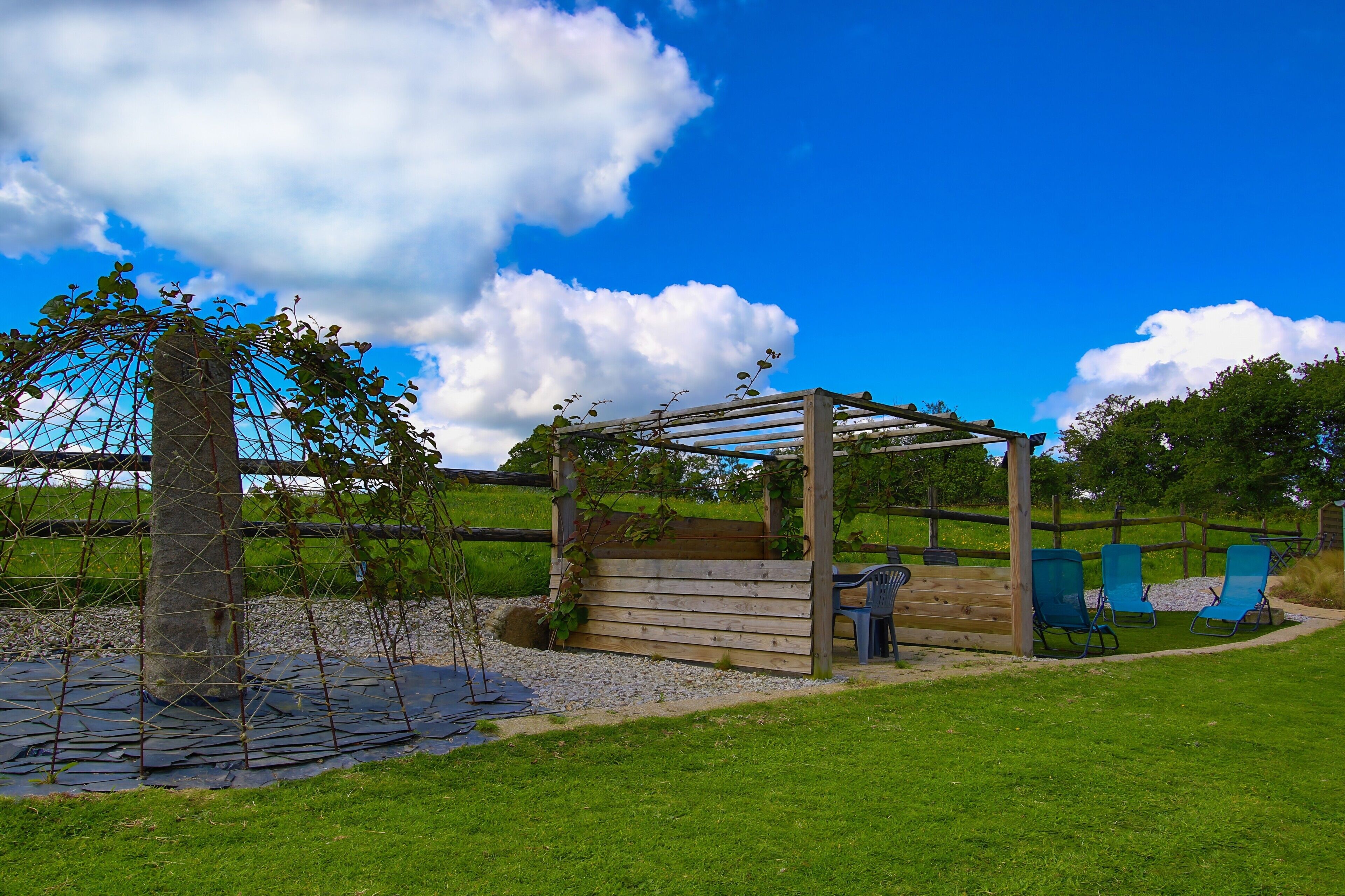 Una pérgola de madera con plantas trepadoras, un pilar de piedra y sillas azules en un área verde al aire libre.