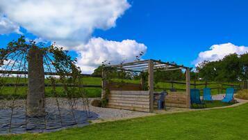 Una pérgola de madera con plantas trepadoras, un pilar de piedra y sillas azules en un área verde al aire libre.