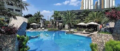 A swimming pool with lounge chairs and umbrellas, surrounded by palm trees and modern buildings.