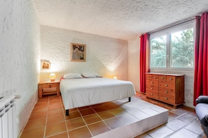 Un dormitori amb un llit, tauletes de nit, un prestatge, una finestra amb cortines i un quadre a la paret.