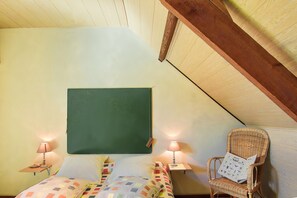 Une pièce comprenant un lit, deux tables de chevet avec des lampes, un tableau noir vert et une chaise en osier avec un coussin.