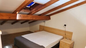 Una camera da letto con soffitto in legno, un letto con materasso bianco, un comodino e una piccola cassettiera in legno.