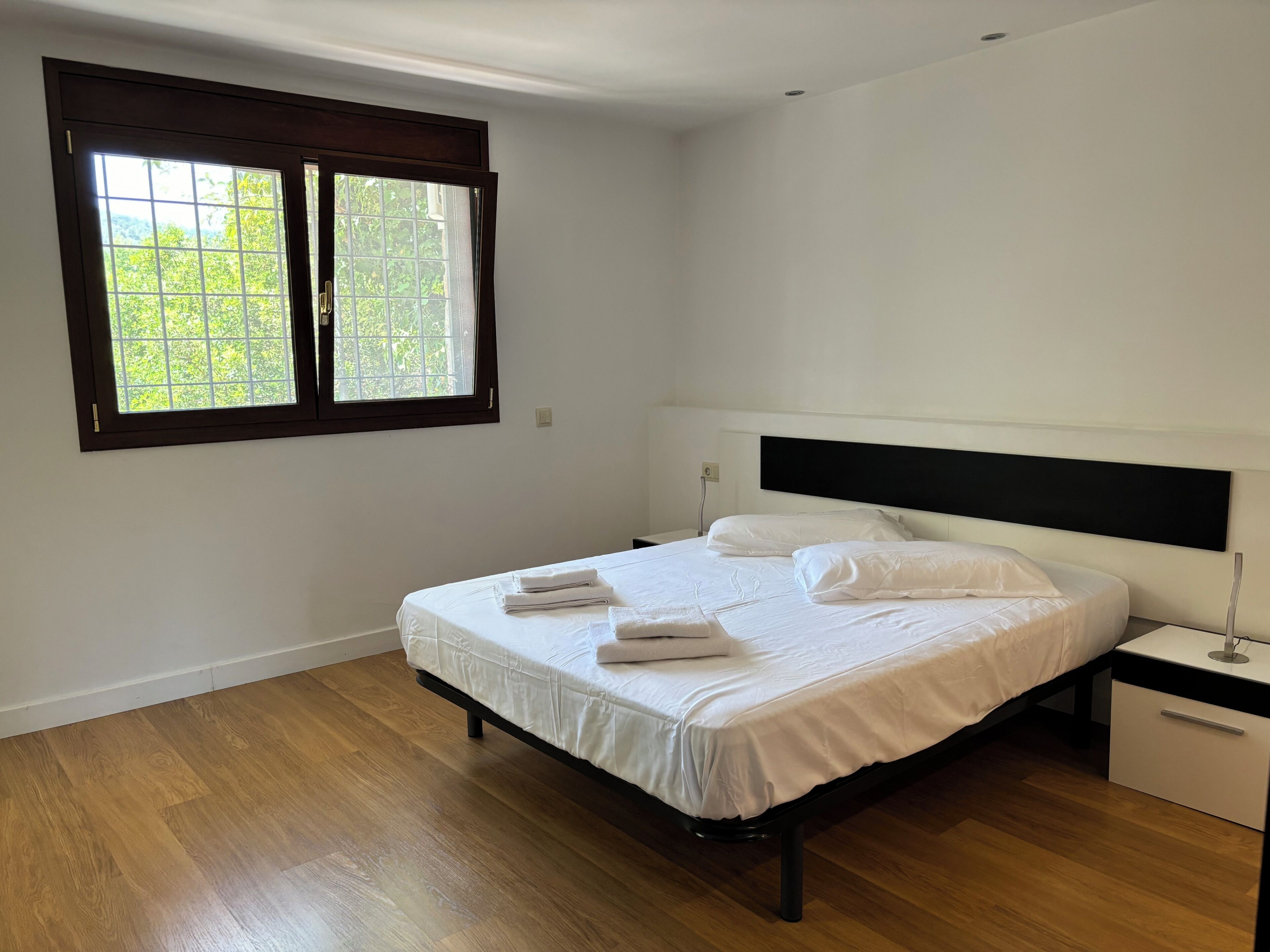 A bedroom with a bed, a nightstand, a window, and a wooden floor.