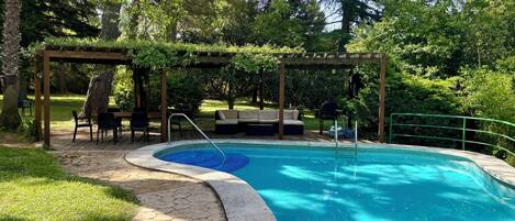 Una piscina con muelle de madera, asientos exteriores y una pérgola.