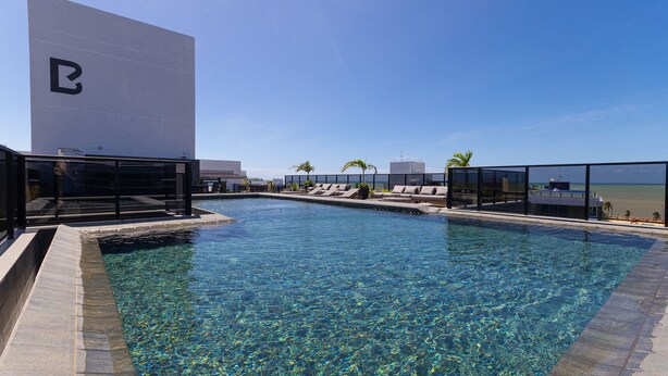 A clear pool with lounge chairs and a large building with a logo. - Way Tambau #R4 - Style and Comfort By Carpediem (João Pessoa)