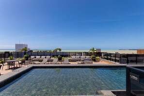 Ein Poolbereich auf dem Dach mit klarem, blauem Wasser, Liegestühlen und Blick auf Strand und Meer.