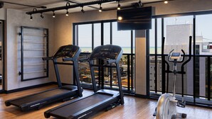 A gym with treadmills, a stationary bike, and a rowing machine. It has large windows with a view of buildings and a body of water. - Way Tambau #R4 - Style and Comfort By Carpediem (João Pessoa)