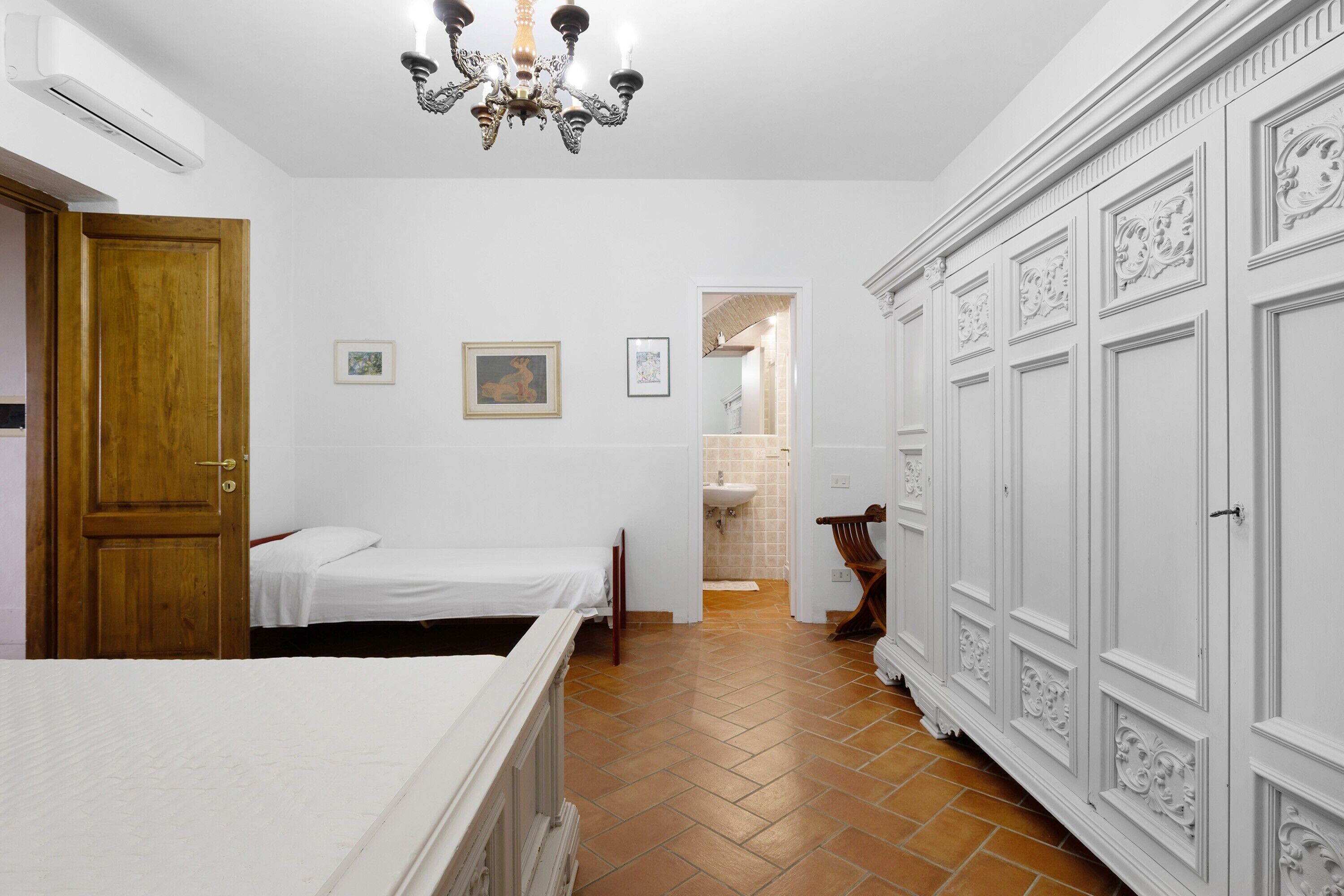 Een slaapkamer met twee bedden, een houten deur, een witte kast, een kroonluchter en een zichtbare badkamer door een open deur.