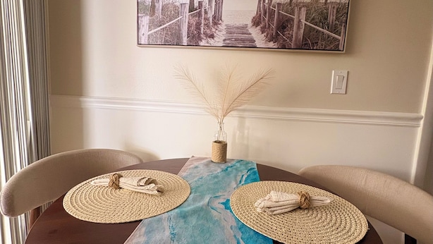 A dining table with two woven placemats, a decorative vase, and a framed beach scene on the wall. - Modern romantic escape designed for comfort. (Jensen Beach)