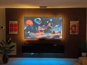 A modern living room with a large flat-screen TV mounted on a wooden wall, a black entertainment unit below, and a potted plant to the left. - Modern romantic escape designed for comfort. (Jensen Beach)