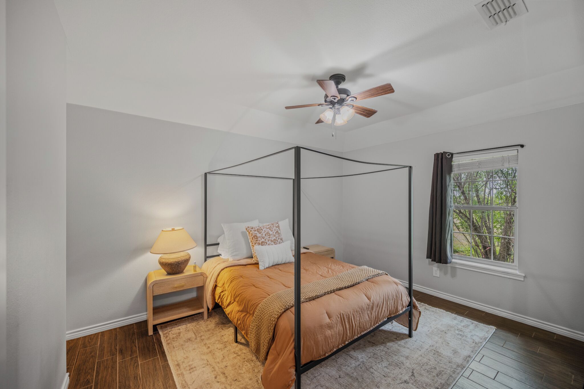 A four-poster bed with a canopy, a bedside table with a lamp, a window with curtains, and a ceiling fan.