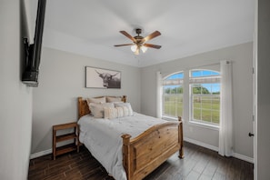 A bedroom with a wooden bed, a ceiling fan, a window with a view of a grassy area, and a mounted TV.