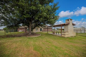 A fenced backyard with a large tree, a stone barbecue area, and a house in the background.