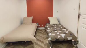 Habitación estrecha con dos camas, una con cubrecama liso y la otra con diseño geométrico, separadas por una pared roja.