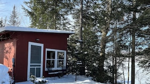 Une cabane rouge aux fenêtres à cadre blanc, entourée d’un paysage enneigé et de grands arbres à feuilles persistantes.