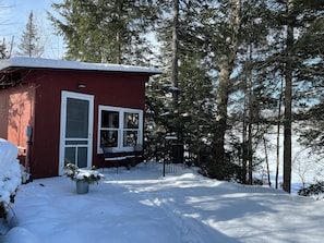 Une cabane rouge aux fenêtres à cadre blanc, entourée d’un paysage enneigé et de grands arbres à feuilles persistantes.
