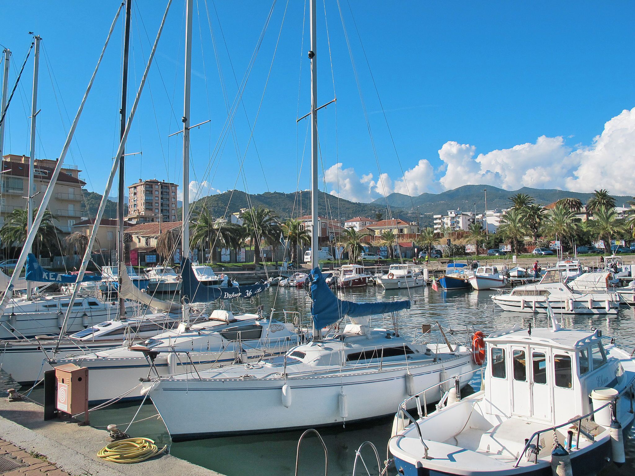 Un puerto deportivo con varias embarcaciones amarradas, cielo despejado y edificios al fondo.