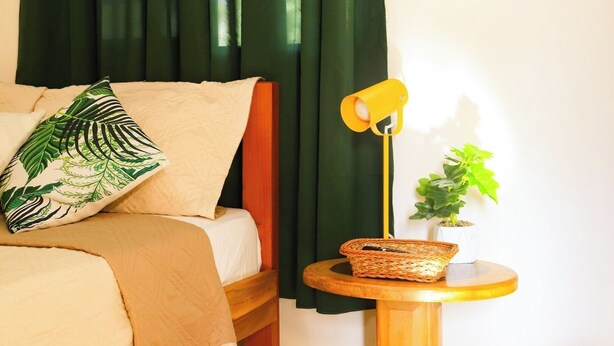 A bed with a green leaf-patterned pillow, a yellow lamp, a wicker basket, and a potted plant on a small wooden table.