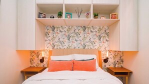 Une chambre à coucher avec un lit, une tête de lit recouverte d’un papier peint à motifs floraux, deux tables de chevet avec des lampes et une étagère intégrée au-dessus du lit.