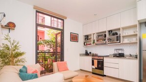 Un espace de vie compact comprenant un coin cuisine, un canapé avec des coussins, une petite table ronde et une vue sur un balcon agrémenté de plantes.