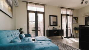 A living room with a blue sofa, a black entertainment unit, and a large window with sheer curtains. - The Glow Stay | Elegant and Modern | @SantaAna (Panama City)