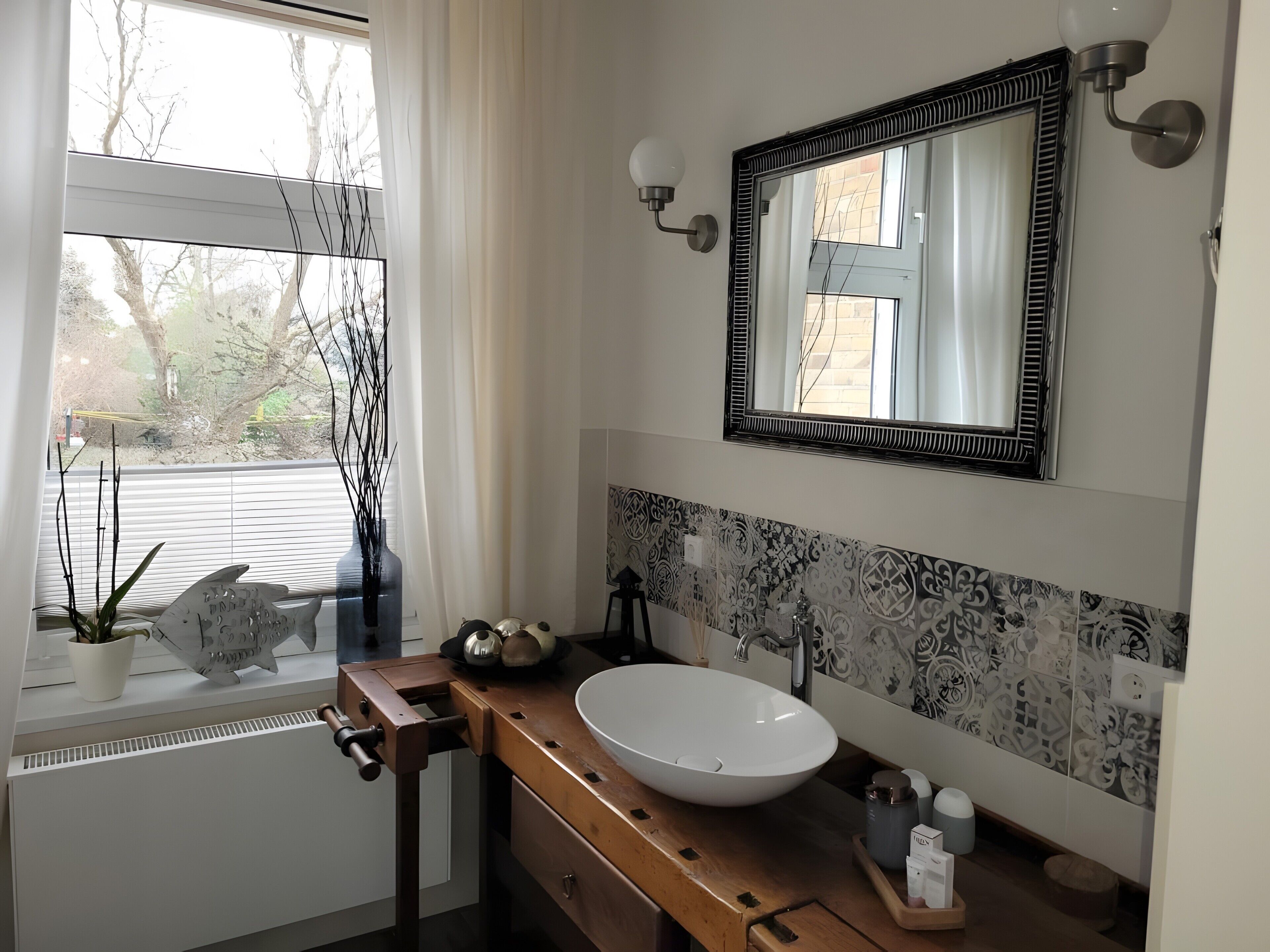 Une salle de bain avec un lavabo blanc, un miroir à cadre noir, une fenêtre donnant sur des arbres et un vase décoratif orné de branches.