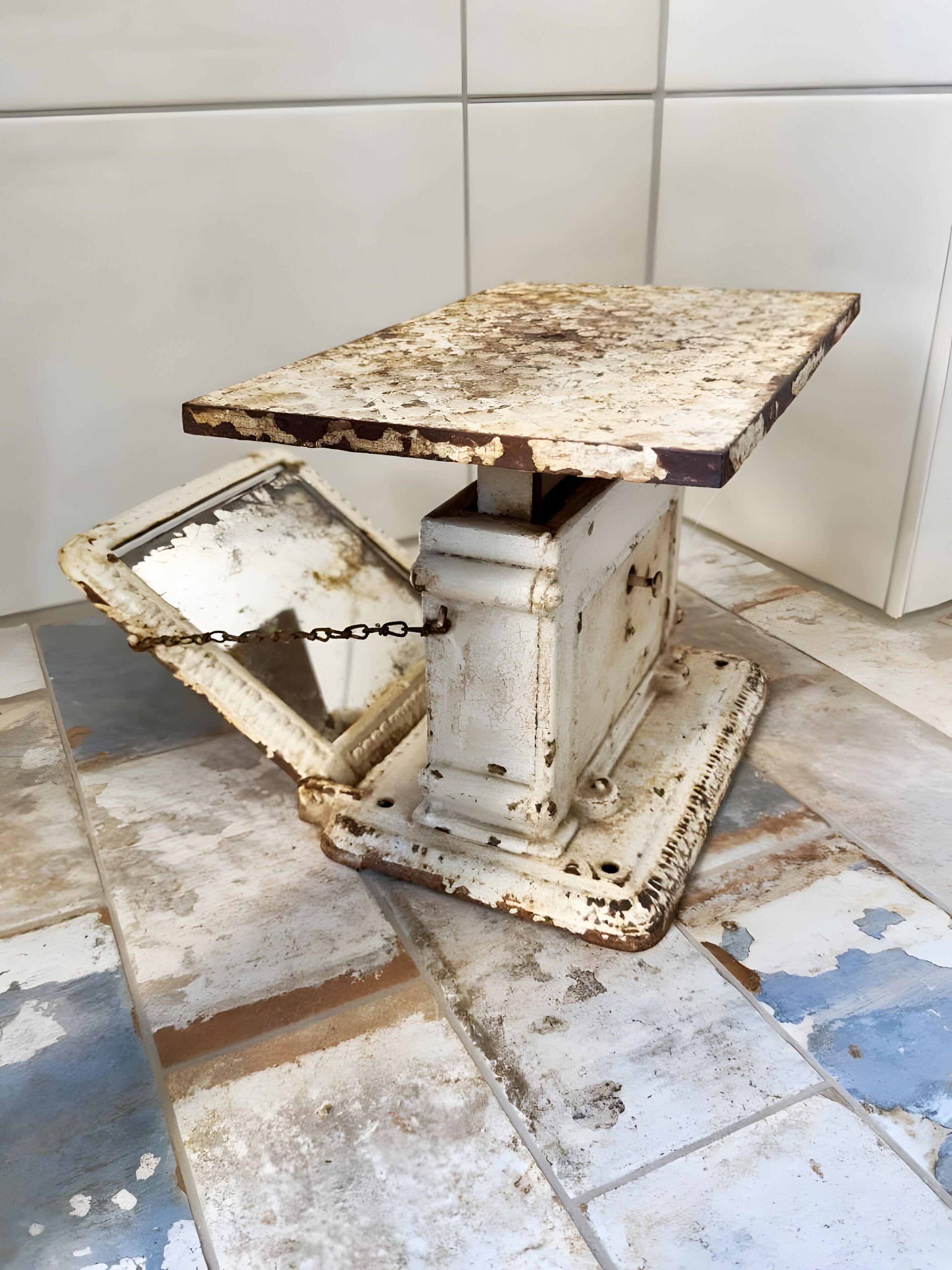 Een rustieke, verweerde tafel met een metalen onderstel en een draaideksel, op een betegelde vloer.