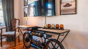 Una stanza con pavimento in legno, una bicicletta, un tavolo con oggetti decorativi, una sedia, un televisore e quadri appesi al muro.