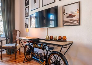 Ein Zimmer mit Holzboden, einem Fahrrad, einem Tisch mit Dekoration, einem Sessel, einem Fernseher und gerahmten Bildern an der Wand.