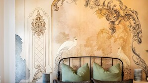 Una camera da letto con un letto, cuscini, un comodino, una cassettiera e un affresco sulla parete.