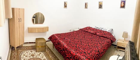 A bedroom with a bed, a wooden wardrobe, a round mirror, a bedside table with a lamp, and a small stool.
