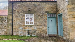 A stone building with a tiled roof, a white window, a blue door, and two metal chairs outside. - The Gardener's Cottage, Low Hall (Scarborough)