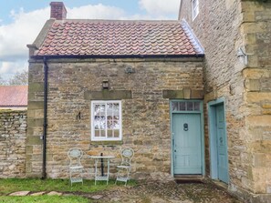 A stone building with a tiled roof, a white window, a blue door, and two metal chairs outside. - The Gardener's Cottage, Low Hall (Scarborough)